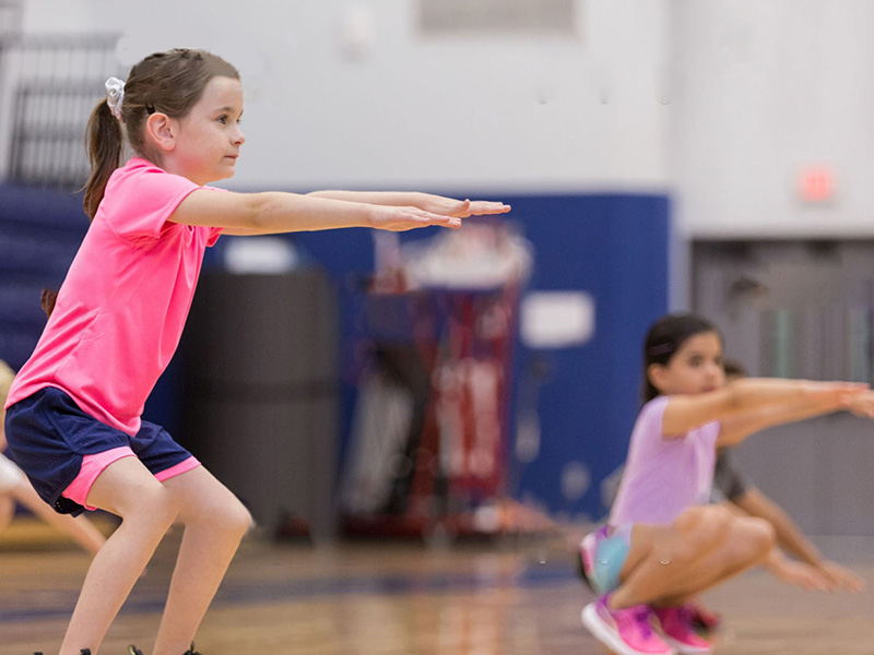 Confident elementary age students do squats during PE class in the school gym.